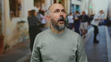 Sakallı kel bir adam, canlı bir şehir ortamında bulanık insanlarla çevrili bir şehir caddesinde şaşırmış bir şekilde duruyor, dışarıda eşsiz ve anlamlı bir an sergiliyor.