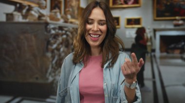 Tablolar ve heykellerle çevrili bir sanat galerisinin içinde gülümseyen İspanyol kadın pembe gömlek ve kot ceket giyerken el hareketleriyle şaşkınlığını dile getiriyor..