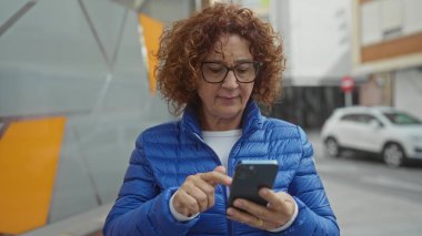 Şehir caddesinde akıllı telefon kullanan kadın gündüz gözlüklü ve mavi ceketli şehir merkezinde park etmiş araçlarla dışarıda günlük aktiviteleri öneriyor.