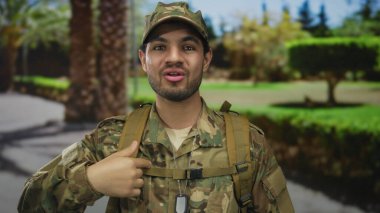 Kamuflaj üniformalı bir adam ve sokakta palmiye ağaçlarıyla el sallayan künye kolyesi; görev ve gurur..