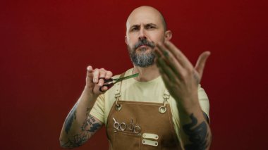 Elinde makas tutan sakallı kel bir adam canlı kırmızı arka plan dövmelerine ve odaklanmış ifadeye karşı önlükle güvenle poz veriyor..