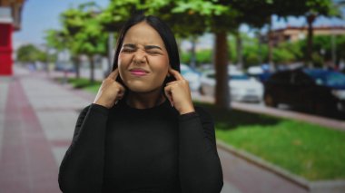 Kadın, kalabalık bir sokakta kulaklarını tutuyor ve Latin kökenli genç adam gürültüden rahatsız oluyor..