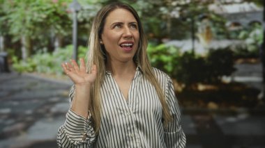 Sarışın, orta yaşlı, gülümseyen ve yeşil yeşillik ve bina cephesiyle el sallayan bir kadın, mutluluğu selamlıyor..