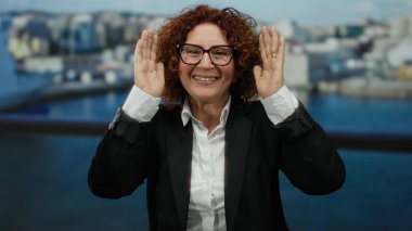 Limanda tekneli ve deniz kenarında gülümseyen bir kadın, esmer, orta yaşlı, İspanyol bir kadın, neşeli bir şekilde yüzünü örtüyor ve mutlu bir ifade sergiliyor..