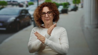 Şehir sokaklarında el kol hareketi yapan bir kadın, gözlüklü orta yaşlı bir İspanyol, beyaz gömlek giyiyor, şehir dışında yaşıyor, arka planda arabalar var, net bir iletişimi vurguluyor..