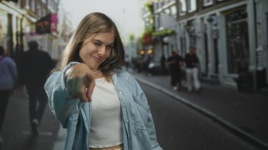 Genç sarışın kadın, işlek şehir caddesinde kamerayı işaret ediyor. Önü açık binalar ve bir bisikletçi. Kendine güveni tam..