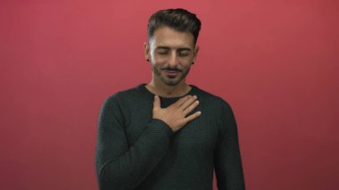 Stüdyoda kırmızı duvara karşı duran koyu renk örülü süveter giyen Latin asıllı genç adam, samimiyet.