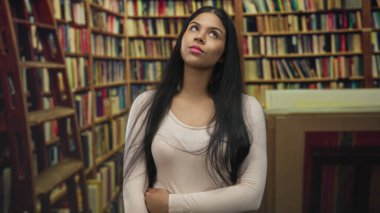 Uzun koyu renk saçlı bir kadın, kitap kaplı binada kollarını kıvırıyor. Düşünceli bakışlı, hafif bir tepe lambası altında..