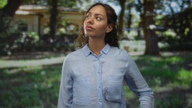 Kadın yüzü parka bakıyor ağaçların yanında ve bir bina, mavi bir gömlek giyiyor; dinginlik tefekkür.