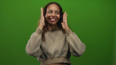 Kırpılmış bir kazak giyerken yüzünü elleriyle çerçeveleyen ve stüdyoda yeşil bir zemine gülümseyen bir kadın; keyif güveni..