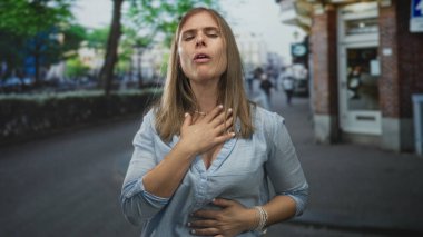 Bir eliyle göğsünü tutan ve diğer eliyle karnını bastıran bir kadın işlek bir şehir caddesinde dikilirken; sıkıntı.