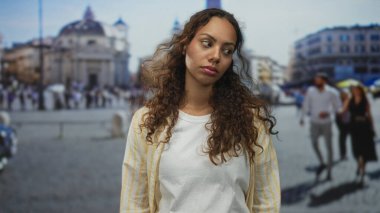 Bir binanın önündeki işlek bir caddede, iki parmağını baş hizasında tutan kadın, gevşek bukleler ve sıradan tişörtler; kayıtsızlık tefekkür.