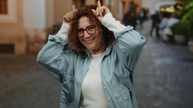 İspanyol orta yaşlı, gözlüklü kadın parmaklarıyla boynuz hareketi yapıyor. Şehrin ortasında neşeli bir ifadeyle duruyor..