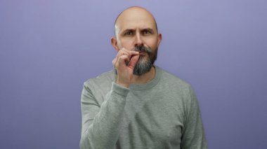 Sakallı yakışıklı kel adam, izole edilmiş mor arka plana karşı gizli bir jest yapar. Gizlilik ya da gizemi ifade eden yüzü ve ince el hareketiyle aktarır..