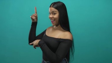 Latin genç bir kadın parmağıyla yukarı ve yana doğru işaret ediyor. Stüdyoda omuzları açıkta ve gülümseyen bir yüzü var..