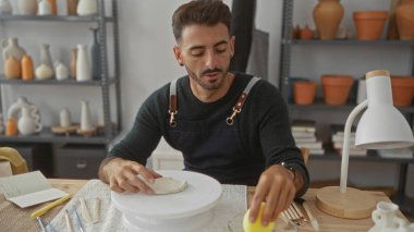 Kapalı mekandaki zanaatkar atölyesinde elleri dönen bir çanak çömlek üzerinde kili şekillendiren adam; dikkatli yaratıcılık.