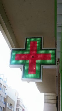 Açık hava ortamında açık mavi gökyüzüne parlayan yeşil ve kırmızı haçlı eczane işareti..