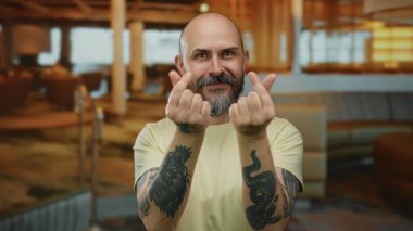 Sakallı, kel bir adam şık bir otelde parmaklarını kalp yapıyor. Kapalı bir odada, arka planda bulanık oturma alanı olan dövmeler yapıyor. Rahat bir ortam yakalıyor.