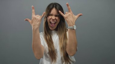 Latin kökenli genç bir kadın, gri bir arkaplan üzerinde iki eliyle neşeli bir rock jesti yapıyor. Beyaz bir gömlekle heyecanı somutlaştırıyor..