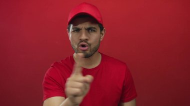Kırmızı şapkalı teslimatçı kaşlarını çatıyor ve kırmızı stüdyoda kameraya işaret ediyor..