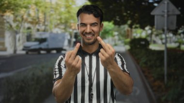 Hakem üniformalı genç İspanyol bir adam şehir sokaklarında bir jest yapıyor ve gülüyor, açık hava ortamında mizah gösteriyor..