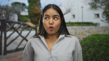 Genç İspanyol bir kadın, güneşli bir günde yeşil bir bahçede komik bir yüz ifadesi takınıyor ve ağaçlar onu kuşatmış..