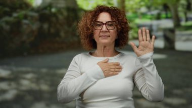 İspanyol kadın, sakin bir park ortamında, gözlük ve beyaz gömlek takarak, yeşil ve taştan yollarla çevrili bir vaadi ya da yemini sembolize ediyor..