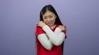 Kırmızı giyinmiş bir kadın mor bir arka plana karşı gözlerini kapatarak kendini kucaklıyor, rahatlatıcı ve barışçıl bir duygu iletiyor, özgüven ve iç huzuru öneriyor..