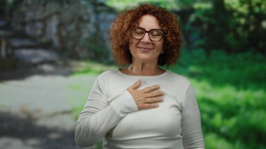 Gözlüklü esmer kadın açık hava parkı ortamında minnettarlığını ifade ediyor. Etrafı yemyeşil..