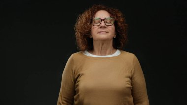 Gözleri kapalı meditasyon yapan bir kadın, turuncu kazaklı esmer, orta yaşlı bir kadın, izole edilmiş siyah duvar arkaplanına karşı, sükunet ve farkındalığı temsil ediyor..