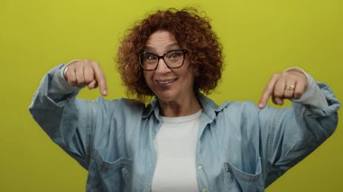 Kıvırcık esmer saçlı, gözlüklü ve mavi gömlekli bir kadın izole edilmiş sarı bir arka planın önünde coşkuyla el kol hareketi yapıyor..