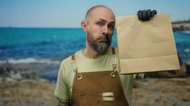 Tulumlu, sakallı bir adam deniz kenarında bir yerde kese kâğıdı tutuyor. Arka planda muhteşem bir deniz manzarası olan düşünceli bir şekilde bakıyor..