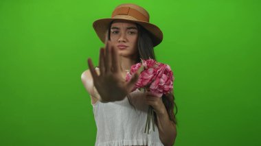 Hasır şapka giyen genç İspanyol kadın yeşil bir stüdyoda el hareketi çekerken elinde pembe bir buket tutuyor..