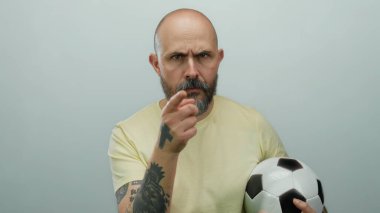 Kel sakallı adam yoğun bir ifadeyle topu beyaz arka planda tutuyor, kolunda dövmeler var..