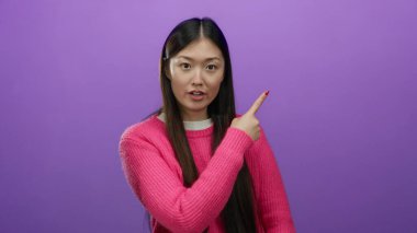 Pembe kazak giyen kadın mor arkaplanı işaret ediyor, canlı renk kontrastını vurguluyor ve izole ortama karşı etkileyici hareketleriyle dikkat çekiyor..