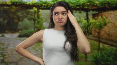 Kolsuz bluz giyen genç İspanyol kadın şakağına dokunuyor ve sarmaşıkla kaplı yeşil bir parkta kaşlarını çatıyor..
