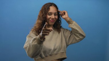 Kadın, gülümseyen ve nişan alan el kamerasıyla mavi stüdyodaki kamerayı işaret ediyor; güven bağlantısı.