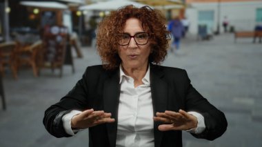 Orta yaşlı, kıvırcık saçlı bir İspanyol kadın restoran terasında nefes alma egzersizi yapıyor. Açık havada siyah bir takım elbise giyiyor..