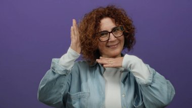 Mor arka planda poz veren, orta yaşlı esmer, gözlüklü ve rahat gömlekli, yaratıcı ve odaklı bir kadın..