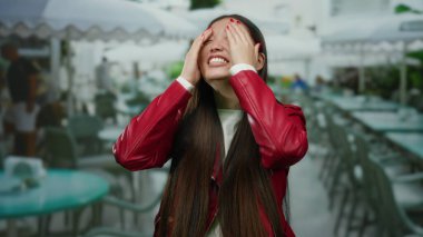 Uzun saçlı ve kırmızı ceketli bir kadın şehir merkezindeki bir terasta neşeyle gülümsüyor, canlı bir restoran ortamının ortasında mutluluk ve tarzını somutlaştırıyor..