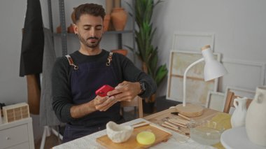 Önlüklü adam atölyedeki çömlek aletleriyle ahşap masadaki kil kasenin yanında kırmızı akıllı telefon dinleme ekranı tutuyor; odaklanmış yaratıcılık.