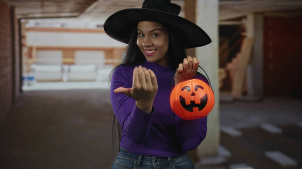 Woman beckons with her hand while holding a pumpkin at an indoor construction building; playfulness.