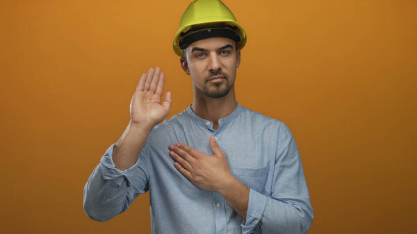 Young man wearing a hardhat making a pledge with one hand on chest and the other raised against an orange background, symbolizing commitment and honesty.