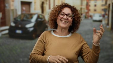 Güneşli bir caddede gitar çalan, gülümseyen ve el kol hareketi yapan bir kadın. Arka planda bulanık arabalar ve tarihi binalar var. Neşeli şehir atmosferini yakalıyor..