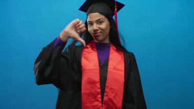 Şapka ve cübbe giymiş genç latin mezunu stüdyoda dudakları büzüşmüş, baş parmak hareketi sergiliyor..