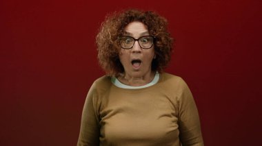 Woman expressing shock in front of a red wall, featuring curly hairstyle, glasses, brown clothing, and isolated interior background.
