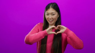 Pembe kazak giymiş, elleri ile kalp şeklinde gülümseyen genç, Asyalı, dışlanmış, dışa dönük, yetişkin, oyuncu bir kadının yer aldığı canlı pembe bir duvara karşı sevgi ve pozitiflik taşıyan bir kadın..