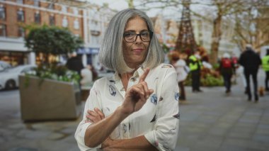 Şehir caddesinde, gün ışığında parmakla işaret eden ve çapraz silah tutan bir kadın, gri saç ve gözlük görünür; kesinlikle onaylanmamış..