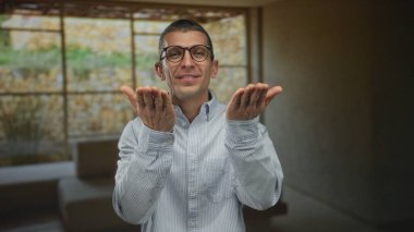 Man indoors in a hotel blowing a kiss wearing glasses and a striped shirt expresses joy in an indoor setting with soft lighting against a blurred window background.