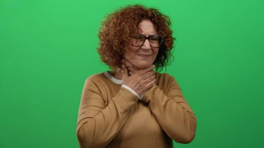 Boğaz ağrısı çeken, rahatsızlık veren ve boynunu tutan, parlak yeşil bir arka plana karşı duran orta yaşlı İspanyol kadının, gözlük takan,.